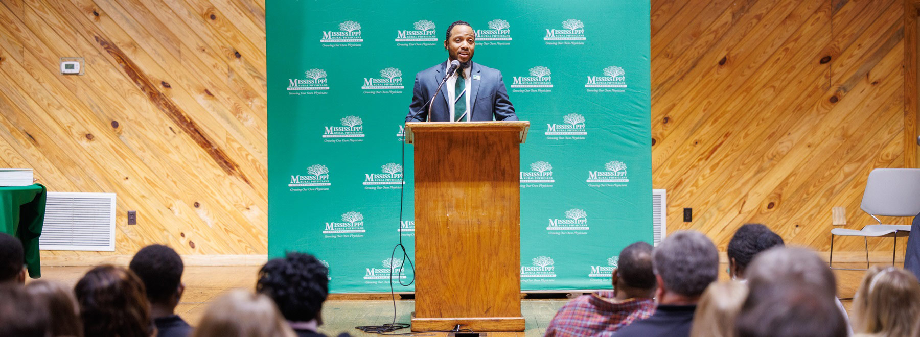 Speaker standing at a wooden podium addressing an audience, with a green Mississippi State University backdrop behind the stage.